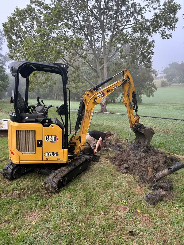 Excavator opening a trench across a lawn to reach a failed drain